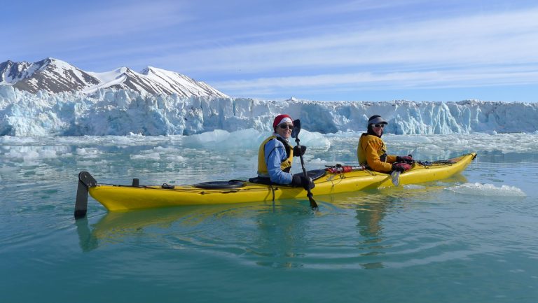 Tandem kayakers near Arctic Circle paddle yellow boat beside glacier ice in glassy turquoise water on a sunny day in Svalbard.