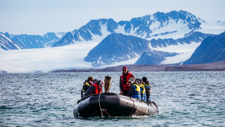 Zodiac boat with Arctic Circle travelers cruises through calm water on a sunny day with snowcapped mountains in the distance.