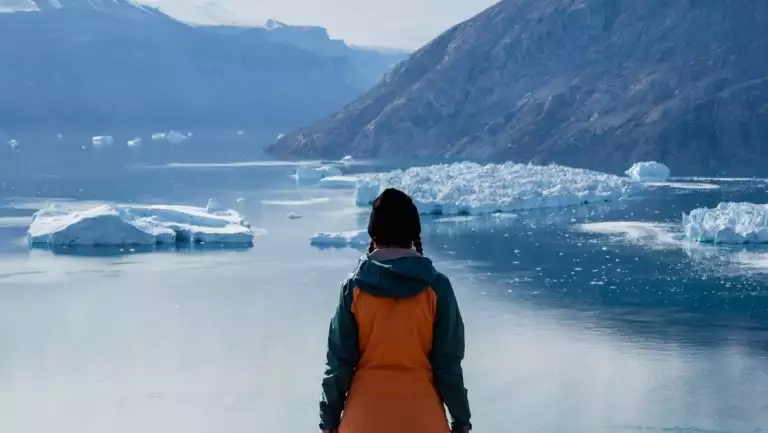 Woman in orange coat & black knit hat stands before glassy fjord with small icebergs during the Arctic Complete cruise.