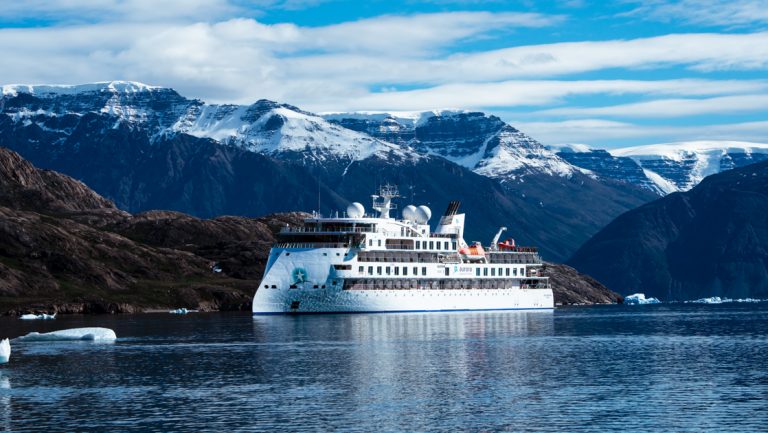 White small expedition ship with pointed bow sits in calm water in a fjord under a partly cloudy & blue sky in the Arctic.