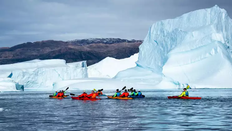 Group of Arctic kayakers with bright jackets paddle red boats in calm water beside large icebergs with brown mountains behind.