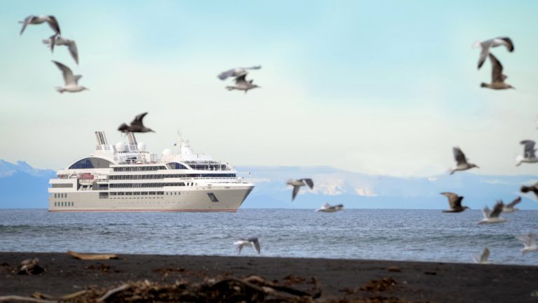 Small ship with gray & red hull cruises past dark shoreline as seagulls fly above during a cruise to Greenland & Baffin Bay.
