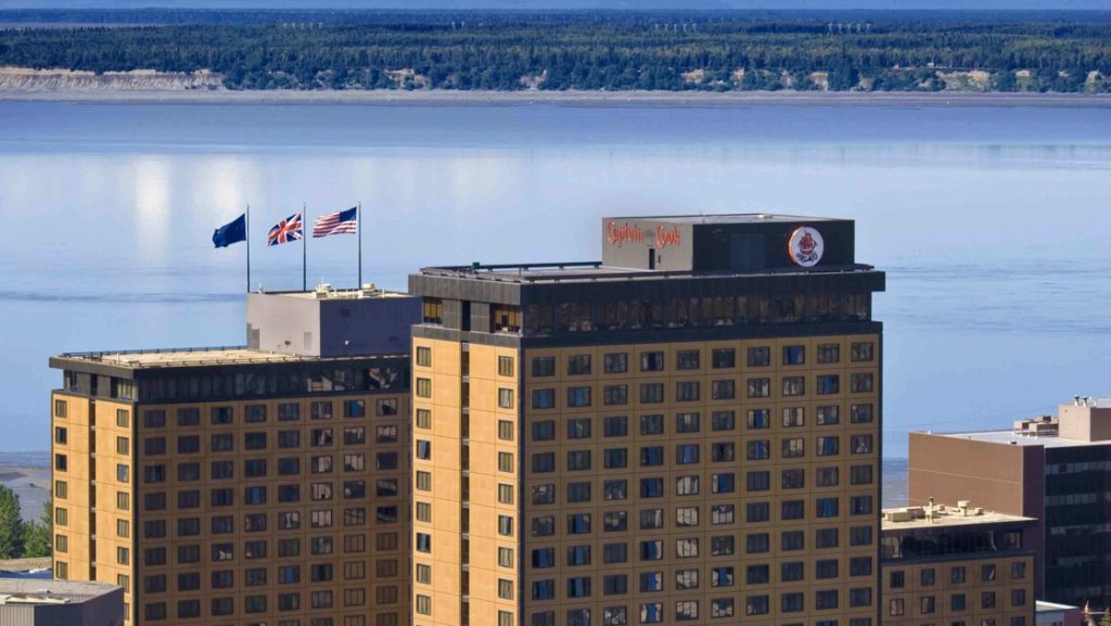 Aerial view of top of Captain Cook Hotel in Anchorage, with 3 beige towers overlooking calm sea with green mountains behind.