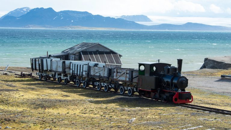 Abandoned train sits on a track among yellow tundra backed by turquoise water with whitecaps & mountains behind, in Svalbard.