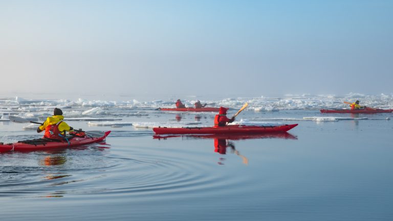 Group of kayakers in red boats paddle glassy water among icebergs on a Commandant Charcot North Pole & Scoresby Sound cruise.