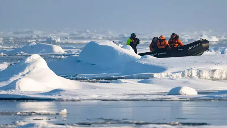 Zodiac boat with North Pole travelers in orange parkas & guide in neon green & black parka sits on an iceberg in the sun.