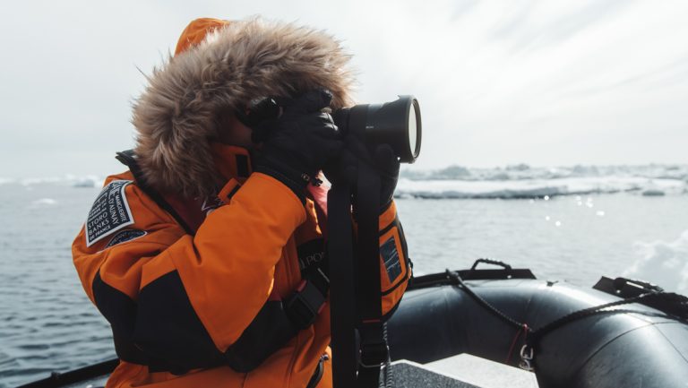 North Pole traveler in orange jacket with fur-lined hood holds camera up to photograph ice while sitting in a Zodiac in the sun.