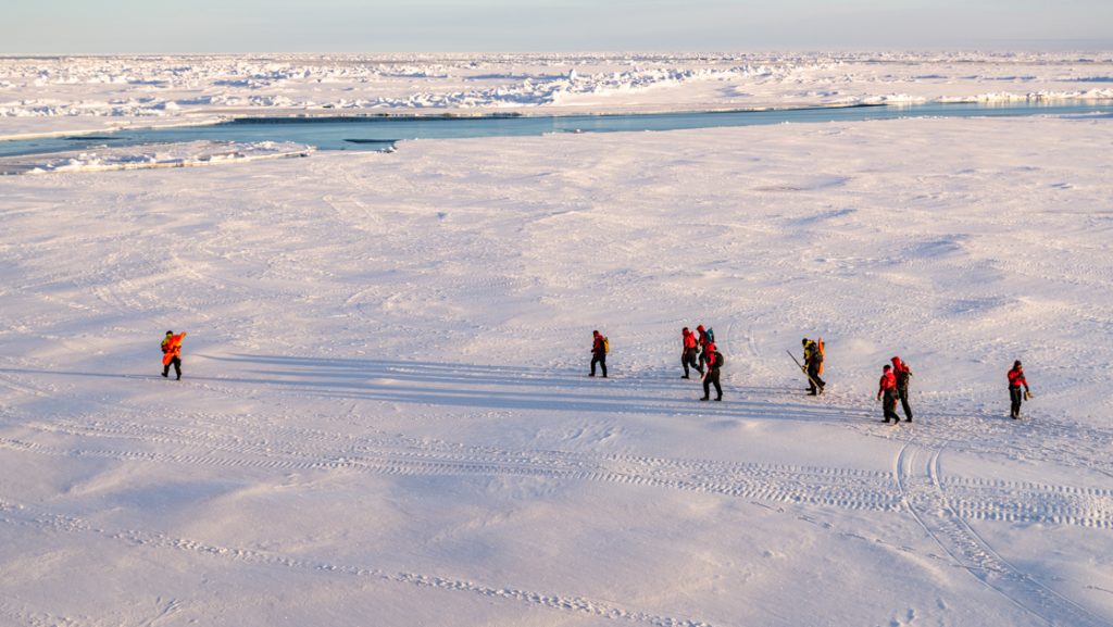 Group of Le Commandant Charcot North Pole travelers in red jackets walks across the ice at dusk, with an ice floe nearby.