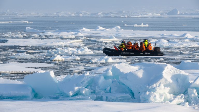 Zodiac full of North Pole travelers in orange jackets cruises among small pieces of ice on a sunny day in the Arctic.