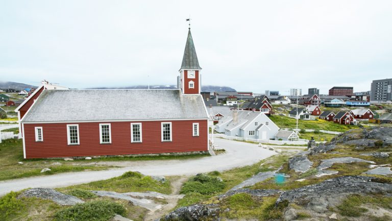 Red church with white trim & gray steeple sits among green & rocky rolling hills & larger town on a cloudy day in Greenland.