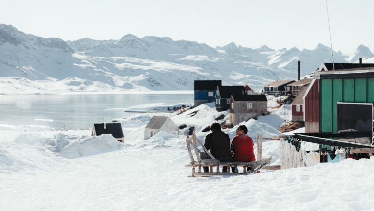 2 men sit on a wooden dog sled in a snow field beside green house with hanging laundry, looking down onto an icy bay in Greenland.
