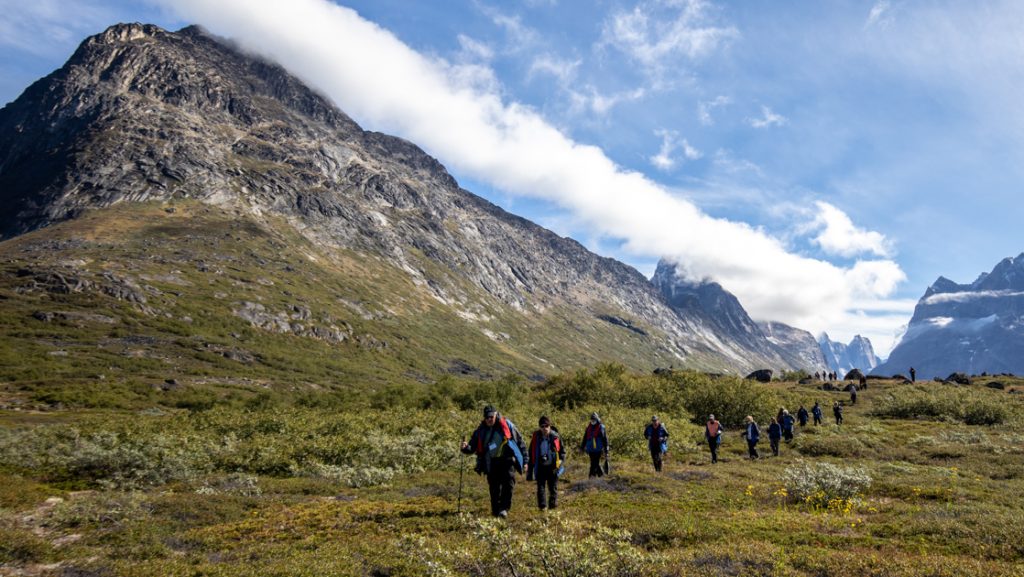 Group of Greenland Odyssey cruise travelers hikes through an alpine valley of green grass under rocky peaks on a sunny day.