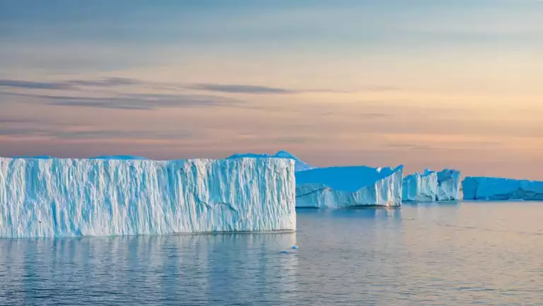 Icy blue tabular icebergs sit in glassy water at sunset, seen on a Greenland Odyssey small ship expedition cruise.
