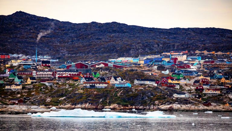 Colorful assortment of small buildings on a seaside town with dark mountains behind, seen on the Greenland Odyssey cruise.