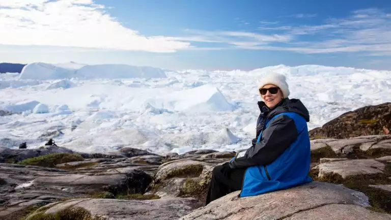 Woman in blue & black jacket & white hat sits on a rock overlooking large glacier covered in snow on a sunny day in Greenland.