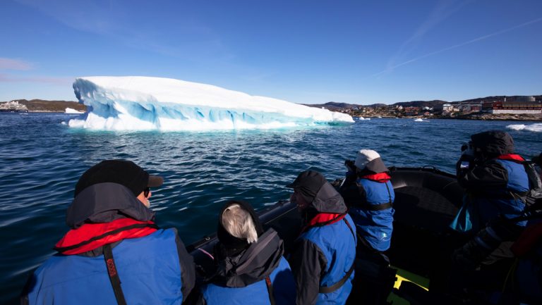 Group of Greenland Odyssey cruise travelers in black & blue coats looks at a large floating iceberg from a Zodiac boat.