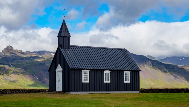 Dark church with white door & trim sits in bright green grass with volcanic mountains behind, under partly cloudy blue sky.