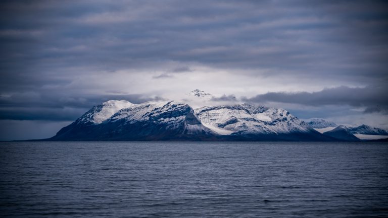 Dark, snow-covered peaks form an island in moderate seas at dusk, seen on an Iceland, Jan Mayen & Svalbard expedition.