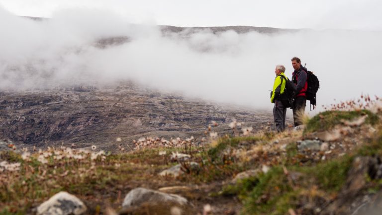 Arctic traveler & guide stand on rocky green tundra by geothermal clouds on a Northern Lights Explorer cruise in the Arctic,