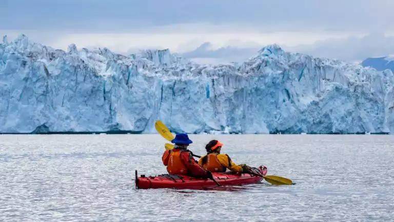 Tandem kayakers in red boat paddle calm water by large blue glacier under cloudy Arctic skies on the Northern Lights Explorer.