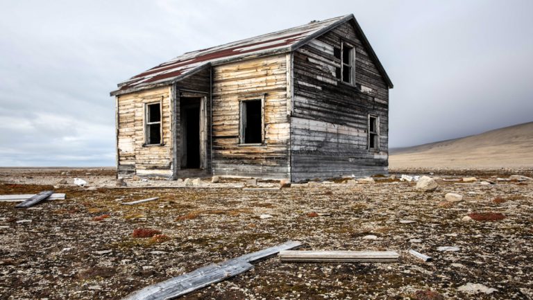 Lone weathered wood hut stands among deserted beige arctic tundra, seen on the Complete Northwest Passage Expedition.