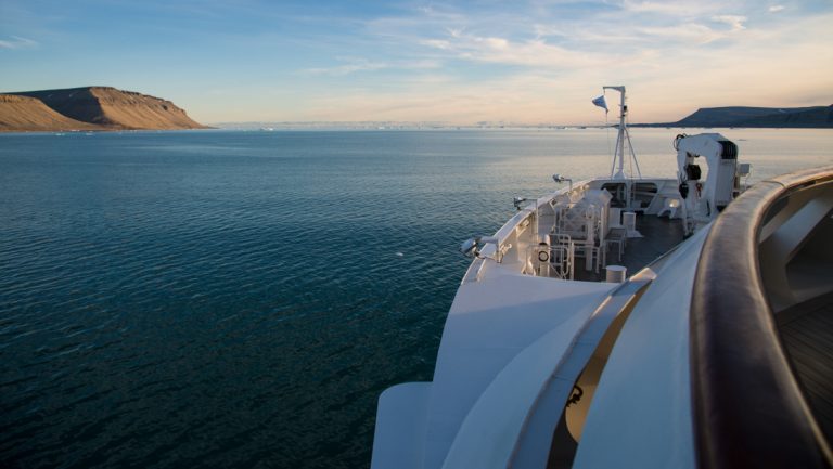View from bow of white ship at sunrise over calm water & tan hills, seen on a luxury Northwest Passage cruise.