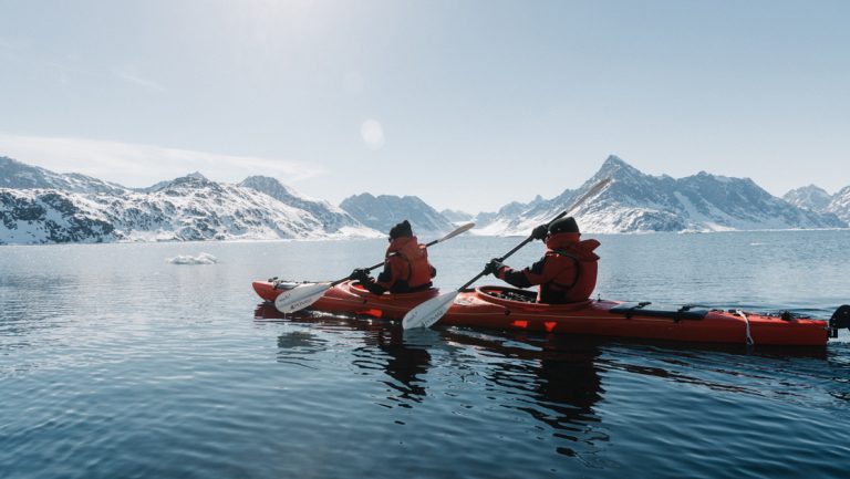 Tandem kayakers in red boat paddle through calm water beside snowcapped peaks on a sunny day of a Northwest Passage cruise.
