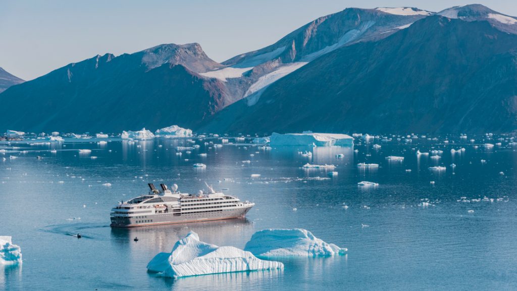 Small expedition ship with gray hull & white upper decks cruises through calm bay among blue icebergs & dark rocky mountains.