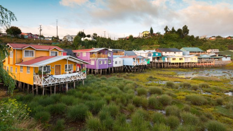 Orange, purple, blue, green, & yellow homes sit on stilts over a marsh, seen during a Patagonia & Chilean Fjords Cruise.