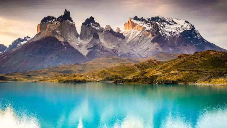 Turquoise lake with golden hillside & jagged peaks behind, seen in Torres del Paine on a Patagonia & Chilean Fjords Cruise.