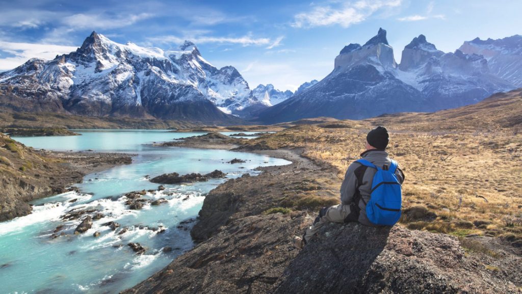 Man in gray parka, black cap & blue backpack sits on a rock by turquoise river, looking up at jagged, snowcapped mountains.