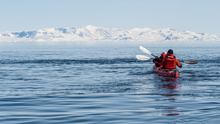 Tandem kayakers in red boat & parkas paddle calm sea beside snow-covered landscapes on a sunny day in Greenland.