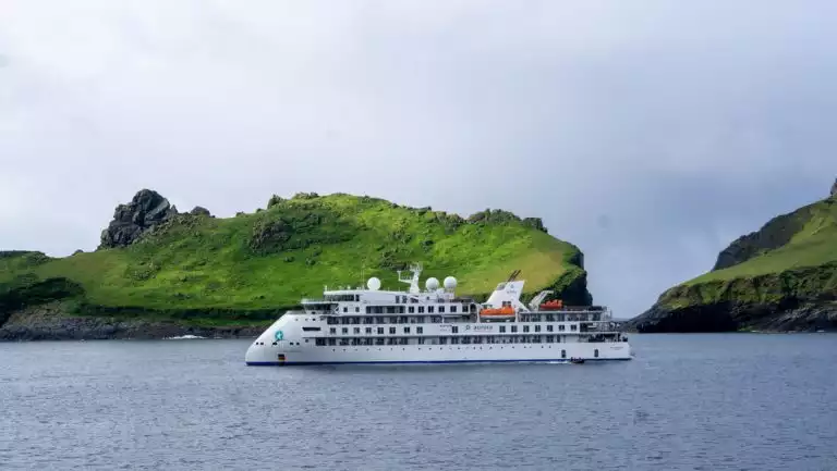 Small white expedition ship with pointed bow sits in calm water by green cliffs on a cloudy day of the Wild Scotland cruise.