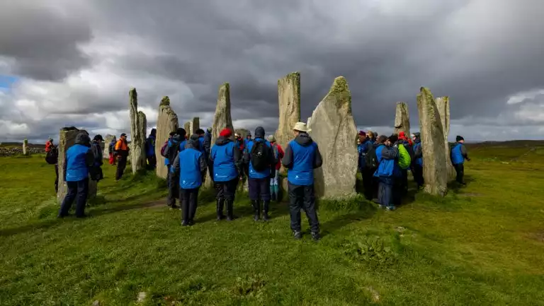 Small group of Wild Scotland cruise travelers in blue jackets walk among primitive tall stone structures in a grassy field.