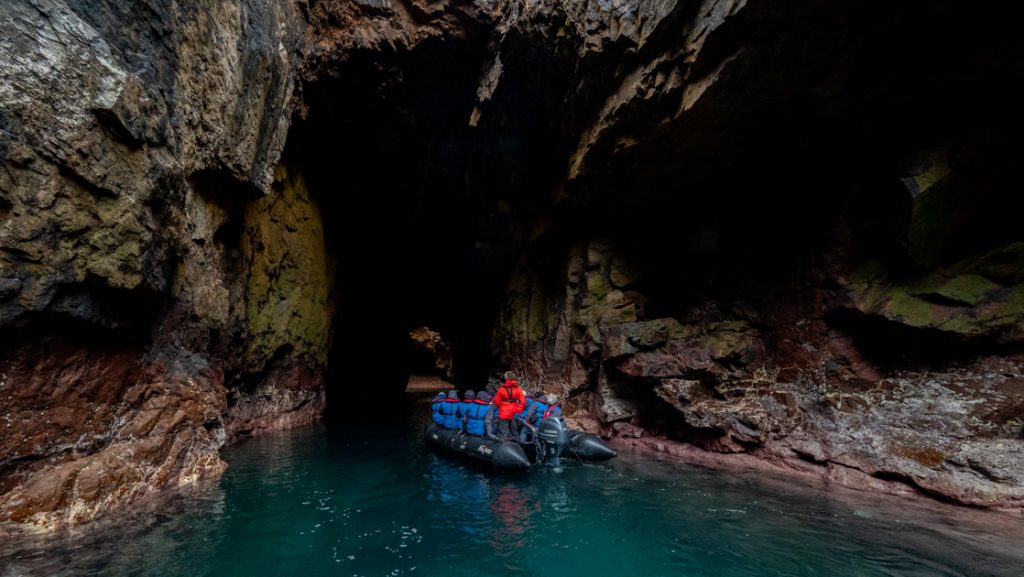Wild Scotland cruise travelers in blue jackets with guide in red parka ride a Zodiac boat into a cave in turquoise water.