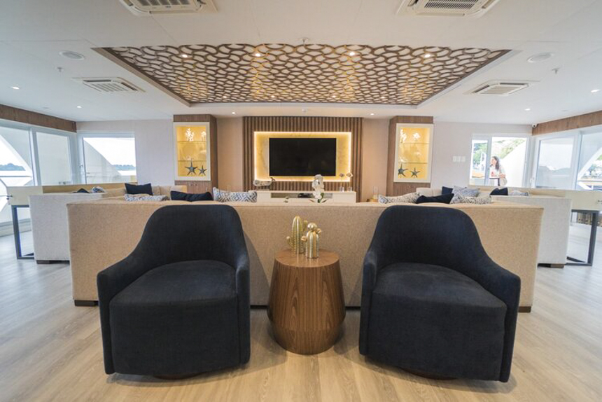 Lounge aboard Elite Galapagos cruise ship, white couches with dark blue accent chairs, flat screen TV and bronze ceiling artwork.