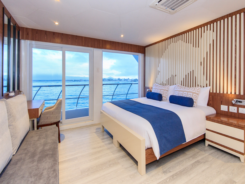 Suite aboard elite Galapagos cruise ship, tortious wood art on wall, white and royal blue bedding, glass sliding doors to balcony