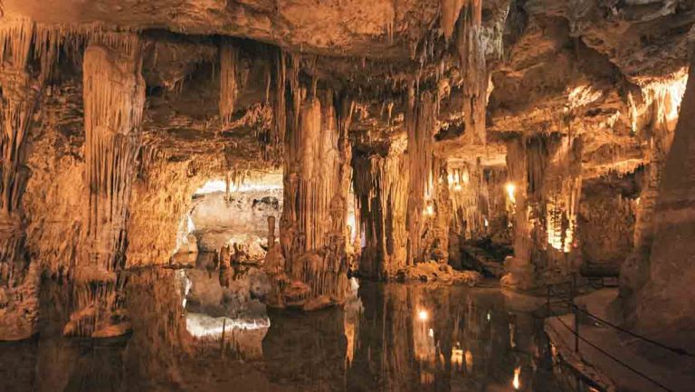 Inside Neptune's grotto in Sardinia, Italy with clear pool of water, tan rock & stalagtites & stalagmites.