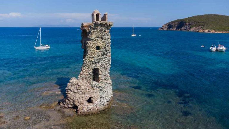 Remains of a medieval building half demolished and flooded by the Mediterranean Sea with small sailboats in the background.