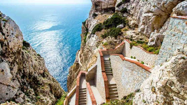 Steep staircase of brick & stone leads up rocky mountainside with turquoise sea below on a sunny day in Sardinia.