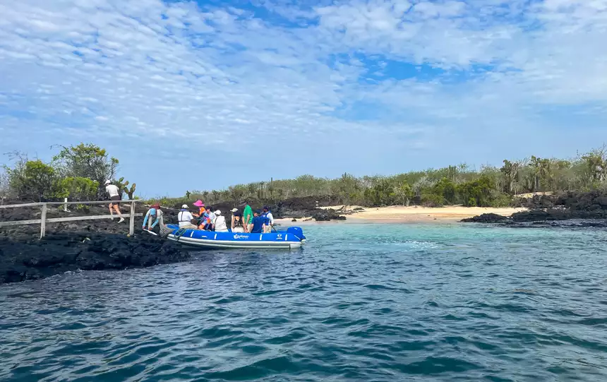 Guests from an Endemic Galapagos cruise exit a royal blue skiff onto the black volcanic rocky shore line on a a sunny day.