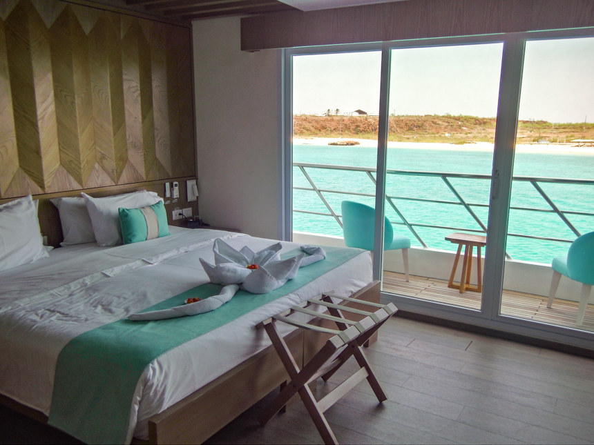Suite aboard Endemic Galapagos cruise ship, with white sheets and teal accents, wood art, and sliding glass doors open to balcony,