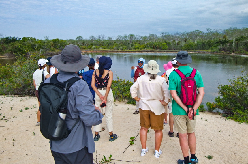 Galapagos Endemic guide stands in front of a brackish lagoon in Galapagos speaking to tour group about flamingos behind them.