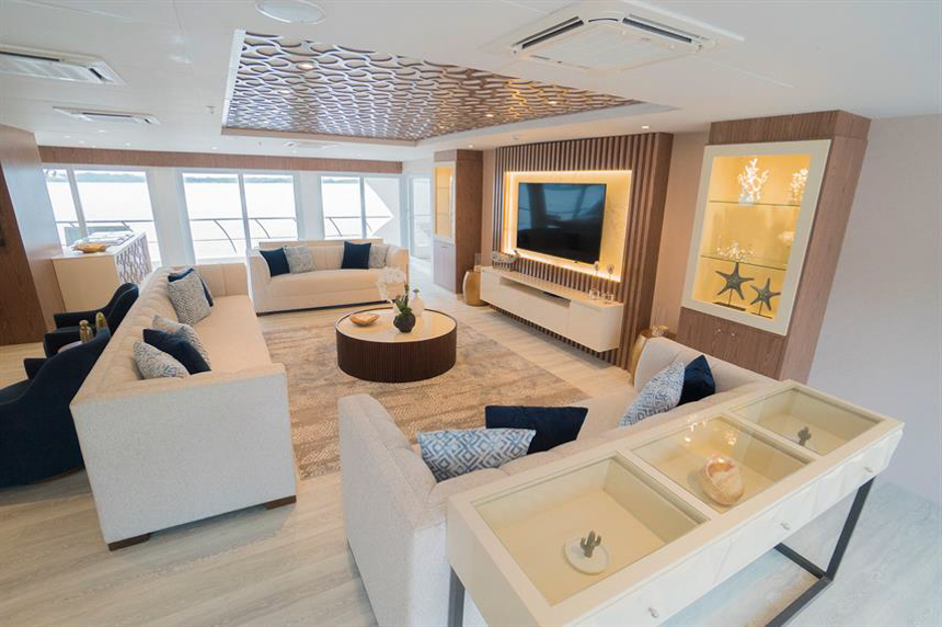 Lounge aboard Elite Galapagos cruise ship, three white couches, round coffee table, dark blue accent chairs, flat screen TV.