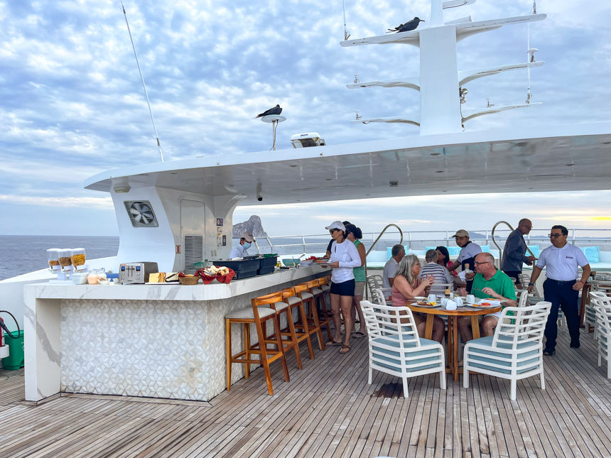 Guests aboard elite Galapagos cruise enjoy lunch at tables on the outside upper deck sun patio.