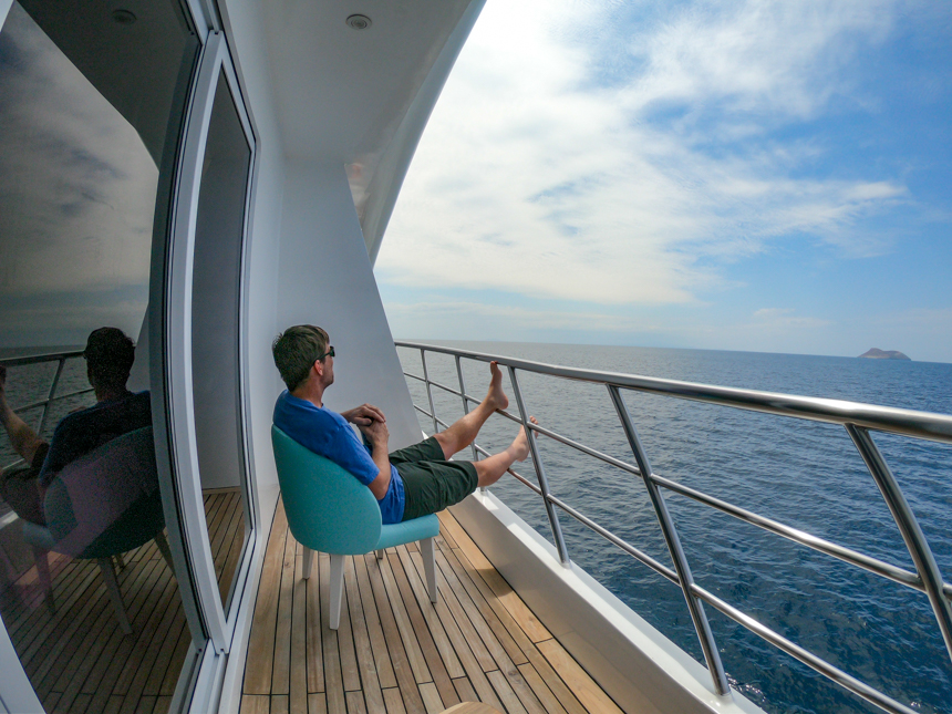 Male traveler sits in teal chair on his private cabin balcony overlooking ocean aboard Elite Galapagos catamaran.