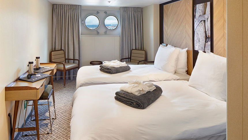 Porthole Cabin on Magellan Explorer with 2 twin beds across from desk with cushioned stool & two chairs, one in each far corner