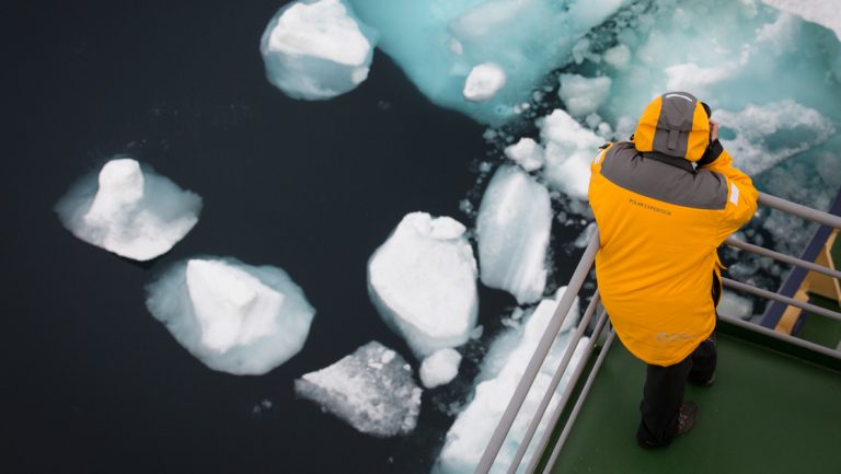Arctic traveler in yellow parka stands at corner of open ship deck, photographing floating bright white & blue iceberg bits.