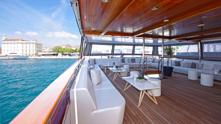 The outdoor covered lounge with white bench couch seating on the Aurora yacht in Croatia with a city shoreline behind it