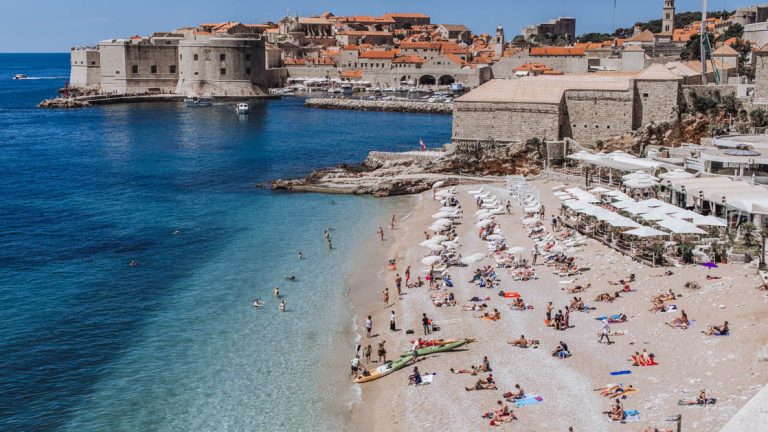 A beach scene in Dubrovnik Croatia seen from above with kayaks on the sandy shoreline and lots of white umbrellas